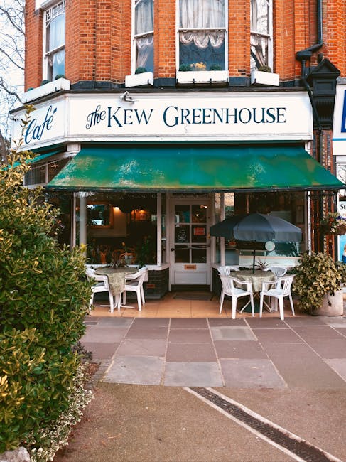 The exterior of 'The Kew Greenhouse' café features a brick building with large sash windows. A white signboard displays the café's name in elegant lettering, supported by a weathered green awning ex