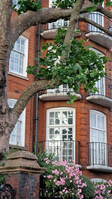 A close-up view of a large, mature tree with textured, grayish-brown bark and broad green leaves partially overshadowing a red-brick residential building in Parsons Green. The building features multip